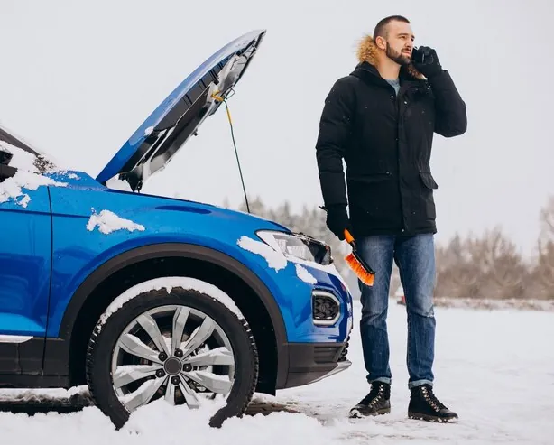 Șofer care sună după ajutor lângă mașina cu capota ridicată, pe zăpadă